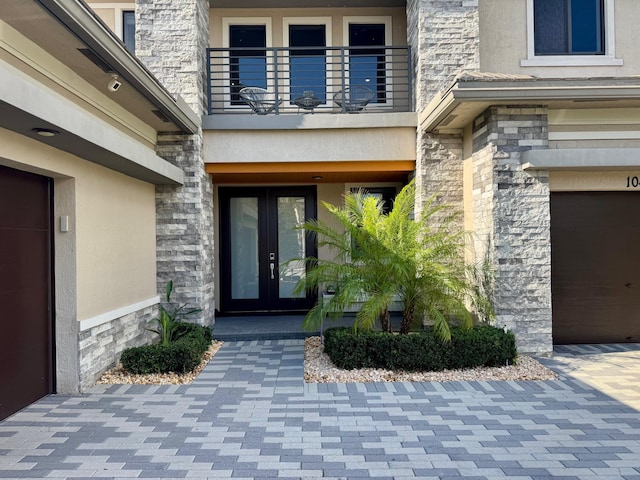 view of exterior entry with french doors, a balcony, an attached garage, stone siding, and stucco siding