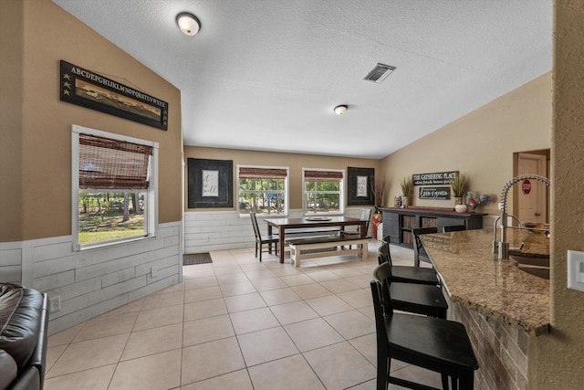 interior space with a wainscoted wall, visible vents, a textured ceiling, light tile patterned floors, and vaulted ceiling