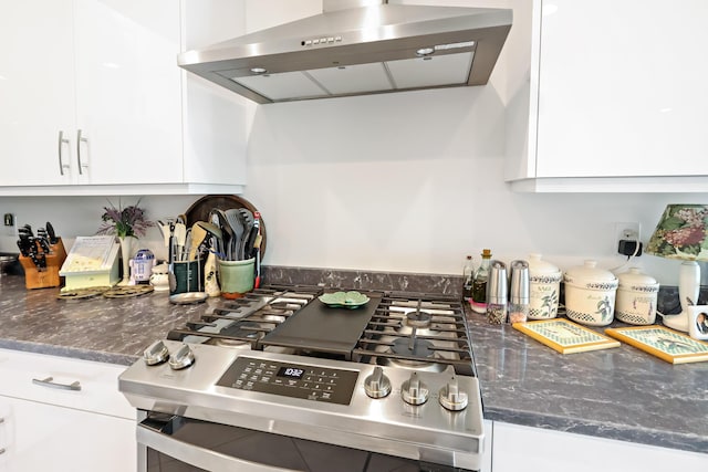 kitchen with dark stone countertops, stainless steel gas stove, white cabinetry, and wall chimney range hood