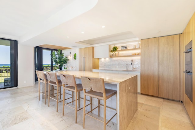 kitchen featuring a sink, backsplash, light countertops, a large island, and open shelves