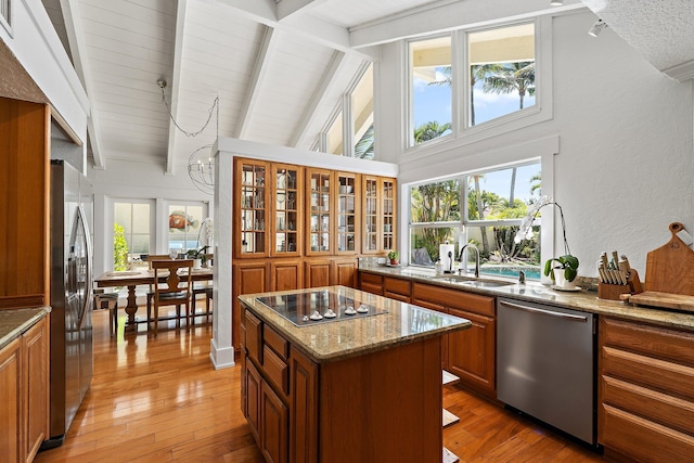 kitchen with beamed ceiling, light stone counters, brown cabinets, appliances with stainless steel finishes, and light wood-style flooring