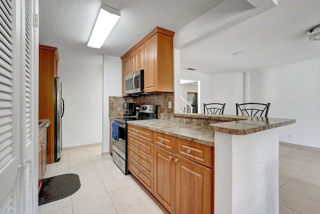 kitchen with a kitchen bar, backsplash, appliances with stainless steel finishes, a peninsula, and light tile patterned floors