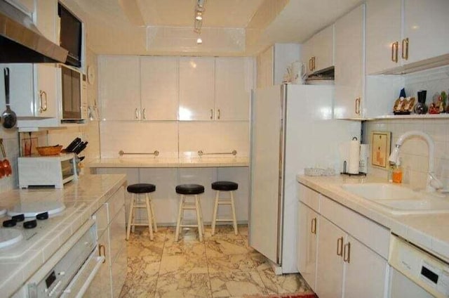 kitchen featuring marble finish floor, a sink, under cabinet range hood, white appliances, and decorative backsplash