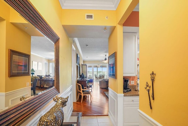 hall with visible vents, a wainscoted wall, ornamental molding, a textured ceiling, and light tile patterned floors