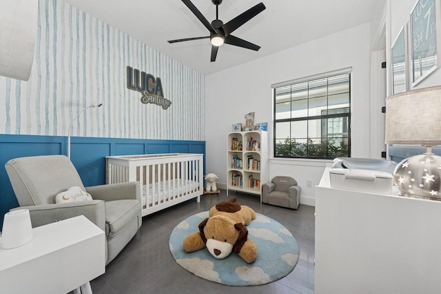 bedroom featuring wallpapered walls, an accent wall, a crib, wainscoting, and a ceiling fan