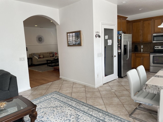 kitchen with light tile patterned floors, brown cabinetry, arched walkways, decorative backsplash, and appliances with stainless steel finishes