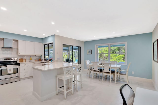 kitchen with wall chimney range hood, stainless steel electric stove, light countertops, a peninsula, and a kitchen breakfast bar