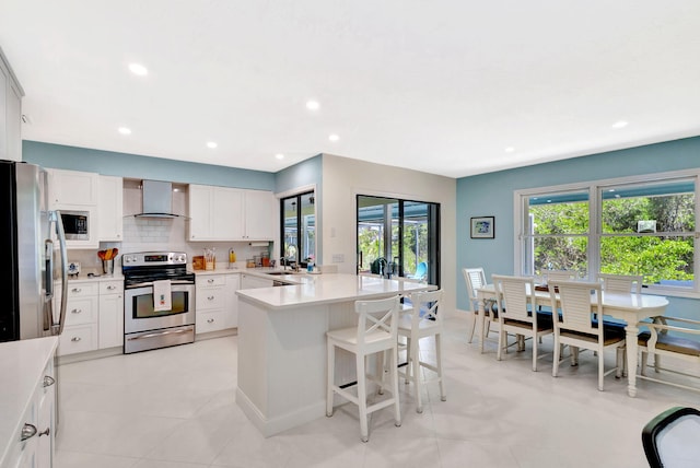 kitchen with a kitchen bar, a sink, stainless steel appliances, a peninsula, and wall chimney range hood