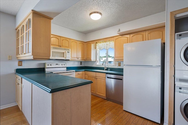 kitchen featuring white appliances, a sink, light brown cabinetry, dark countertops, and stacked washer / dryer