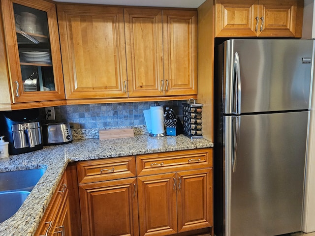 kitchen featuring light stone counters, brown cabinetry, freestanding refrigerator, glass insert cabinets, and backsplash