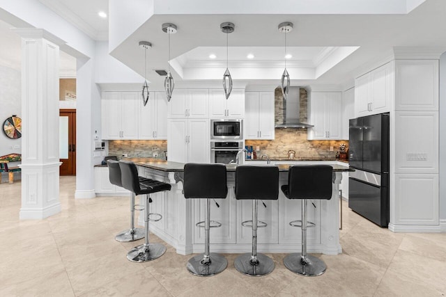 kitchen with crown molding, wall chimney range hood, a tray ceiling, decorative columns, and appliances with stainless steel finishes