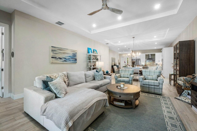 living area with visible vents, a raised ceiling, light wood-style flooring, and ceiling fan with notable chandelier