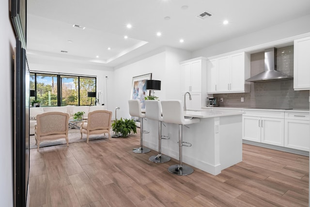 kitchen with tasteful backsplash, wall chimney range hood, a center island with sink, light countertops, and light wood-type flooring