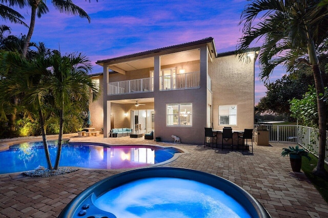 back of property at dusk featuring a balcony, a patio, an in ground hot tub, stucco siding, and ceiling fan