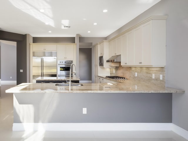 kitchen with backsplash, light stone countertops, under cabinet range hood, built in appliances, and a peninsula