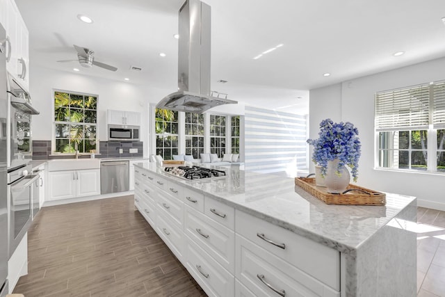 kitchen with stainless steel appliances, white cabinetry, a ceiling fan, light stone countertops, and island range hood