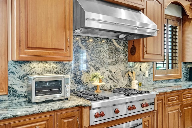 kitchen featuring wall chimney exhaust hood, light stone countertops, stainless steel gas cooktop, decorative backsplash, and a toaster