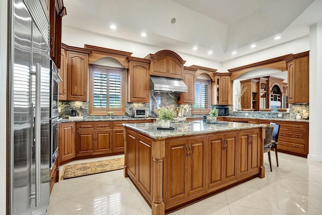 kitchen with light tile patterned floors, a center island, exhaust hood, built in refrigerator, and brown cabinetry