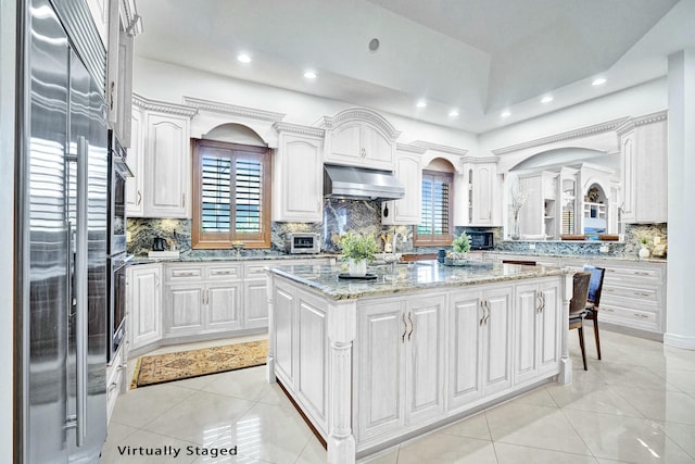kitchen with white cabinets, stainless steel built in fridge, under cabinet range hood, a kitchen island, and light tile patterned flooring