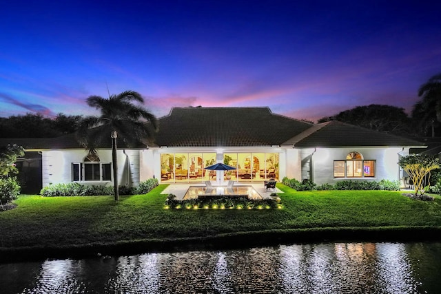 back of house at dusk featuring a patio, a lawn, stucco siding, and a water view