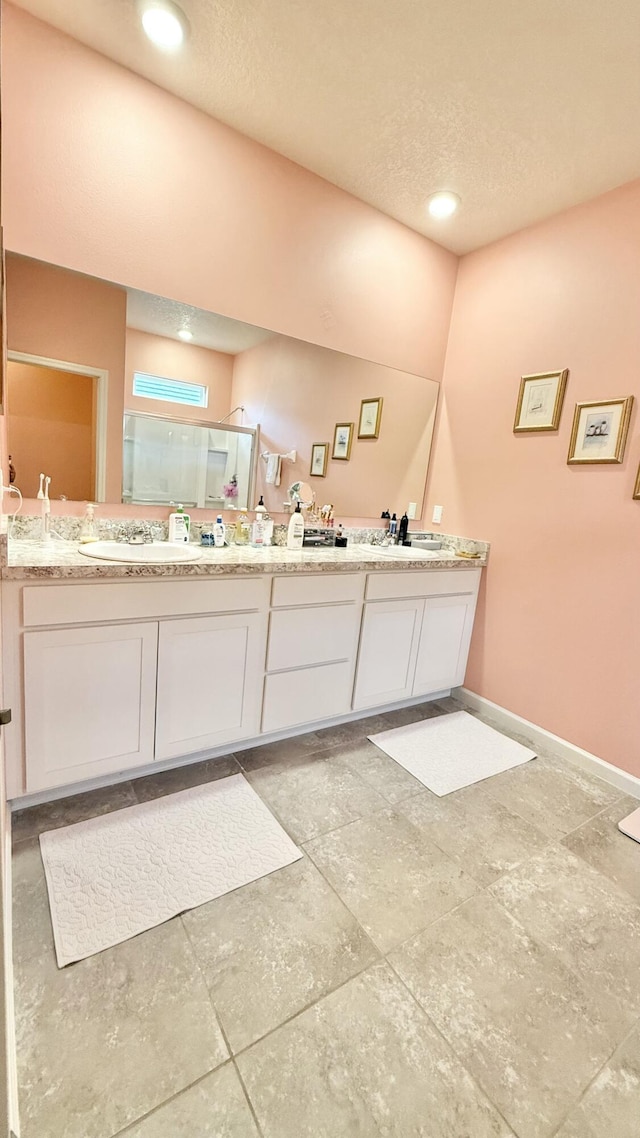 full bath featuring a textured ceiling, baseboards, double vanity, a sink, and a stall shower
