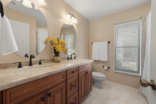 full bath with toilet, a sink, double vanity, and baseboards