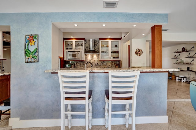 kitchen with stainless steel microwave, a kitchen breakfast bar, wall chimney range hood, glass insert cabinets, and visible vents