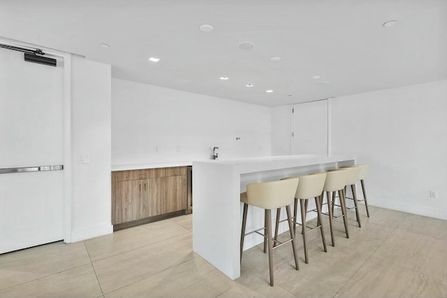 kitchen featuring light countertops, a kitchen breakfast bar, brown cabinetry, baseboards, and recessed lighting