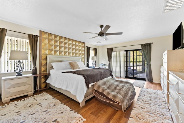 bedroom with ceiling fan, access to exterior, visible vents, an accent wall, and wood finished floors