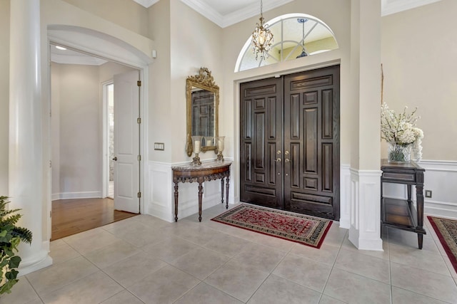 entrance foyer with crown molding, ornate columns, light tile patterned flooring, a decorative wall, and wainscoting