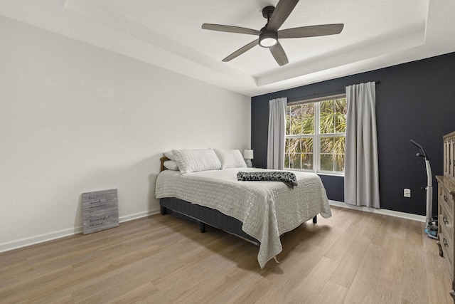 bedroom with a tray ceiling, light wood-type flooring, a ceiling fan, and baseboards
