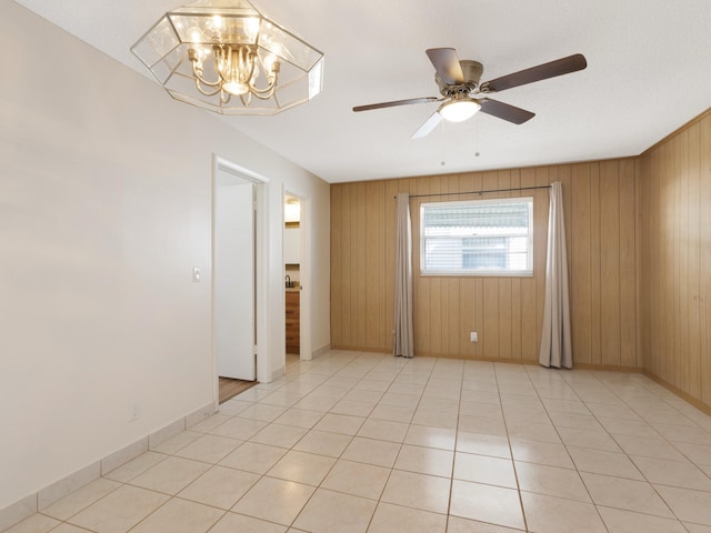 empty room featuring baseboards, wooden walls, light tile patterned floors, and ceiling fan with notable chandelier