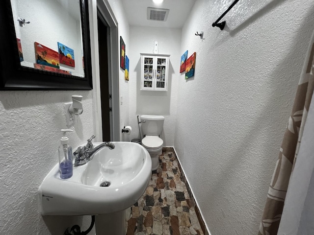 bathroom with stone flooring, baseboards, toilet, a textured wall, and a sink