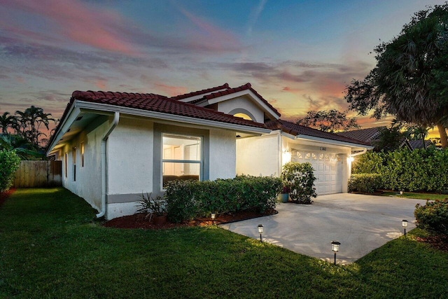 property exterior at dusk with a tile roof, stucco siding, fence, an attached garage, and a lawn