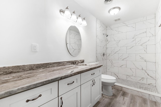 full bathroom featuring a tile shower, toilet, vanity, wood finished floors, and visible vents
