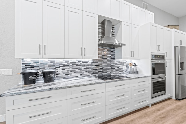 kitchen featuring light stone counters, wall chimney range hood, light wood-style flooring, backsplash, and appliances with stainless steel finishes