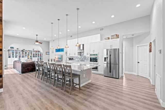 kitchen featuring wall chimney range hood, light wood-style floors, ceiling fan, stainless steel appliances, and arched walkways