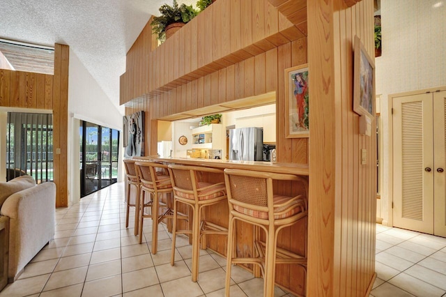 13179 Camero Way, Palm Beach Gardens, FL 33418 - bar with stainless steel fridge with ice dispenser, light tile patterned flooring, wood walls, high vaulted ceiling, and a textured ceiling
