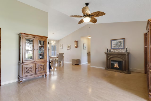 living room featuring high vaulted ceiling, ceiling fan with notable chandelier, a glass covered fireplace, light wood-style floors, and baseboards