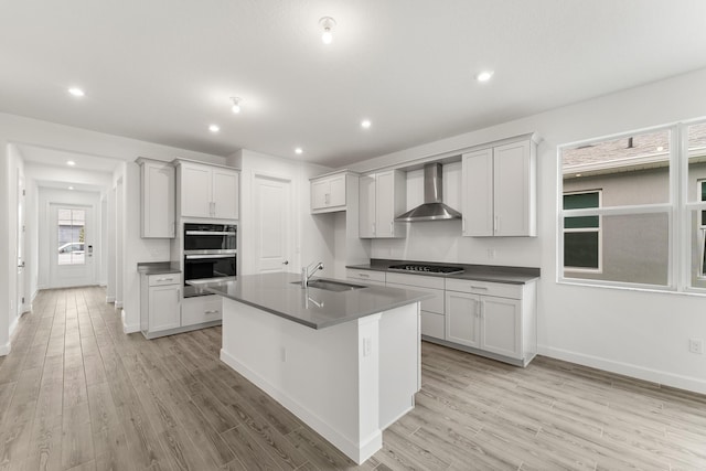 kitchen with a sink, light wood finished floors, stainless steel double oven, wall chimney range hood, and dark countertops