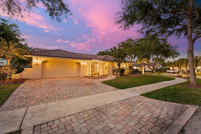 mediterranean / spanish home featuring decorative driveway, a tiled roof, an attached garage, a yard, and stucco siding
