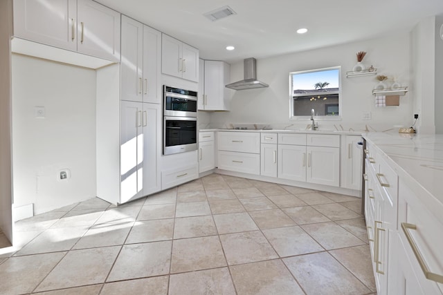 kitchen featuring white cabinets, stainless steel double oven, light stone counters, wall chimney range hood, and visible vents