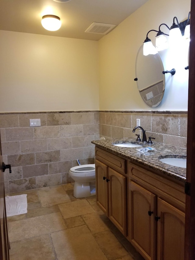 bathroom featuring double vanity, stone tile flooring, visible vents, wainscoting, and a sink