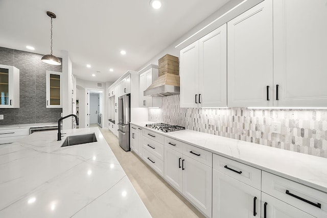 kitchen featuring wall chimney range hood, a sink, glass insert cabinets, stainless steel appliances, and white cabinets