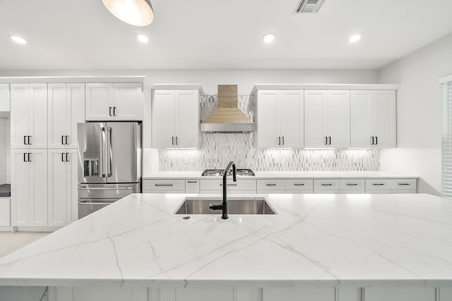 kitchen with wall chimney range hood, a sink, an island with sink, appliances with stainless steel finishes, and visible vents