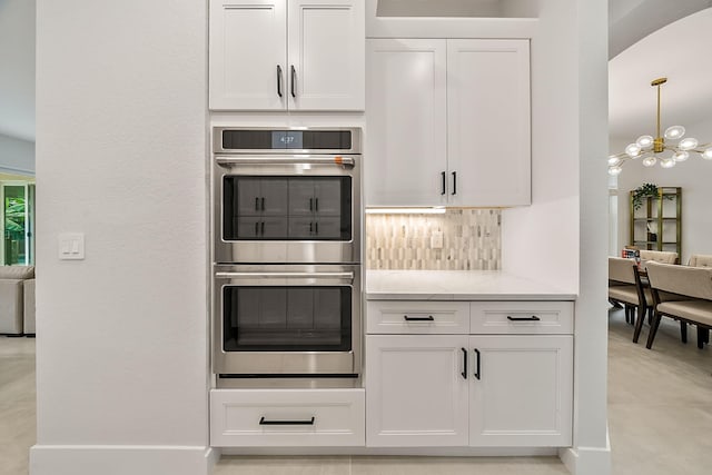 kitchen with light stone countertops, stainless steel double oven, a chandelier, backsplash, and white cabinets