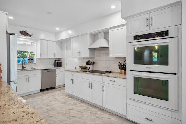 kitchen featuring wall chimney range hood, appliances with stainless steel finishes, a sink, white cabinetry, and backsplash