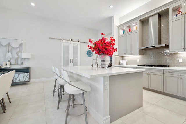kitchen featuring tasteful backsplash, a center island with sink, wall chimney range hood, and light countertops