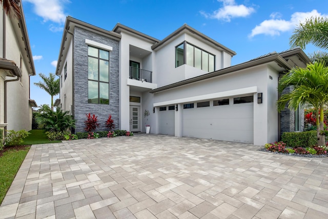 contemporary home with stone siding, a balcony, a garage, stucco siding, and decorative driveway