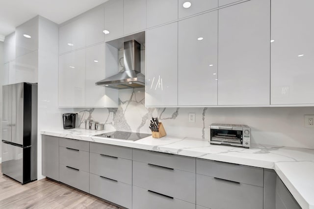 kitchen featuring wall chimney range hood, freestanding refrigerator, black electric stovetop, and gray cabinets